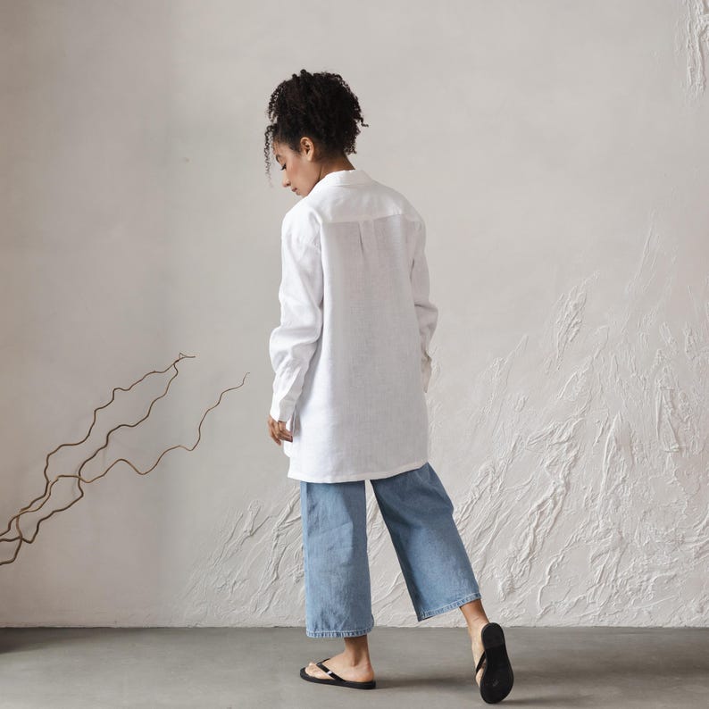 May include: A white linen shirt with long sleeves and a collared neckline. The shirt is loose-fitting and falls to the mid-thigh. The model is wearing blue denim pants and black sandals.