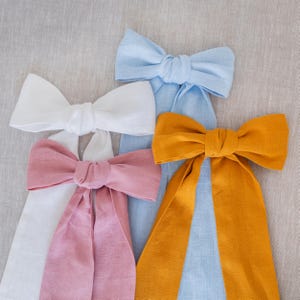 Puede incluir: Cuatro lazos de lino en colores pastel: blanco, rosa, azul claro y amarillo dorado. Cada lazo tiene largas colas fluidas. Los lazos están dispuestos sobre un fondo neutro y texturizado, creando una estética suave y elegante.