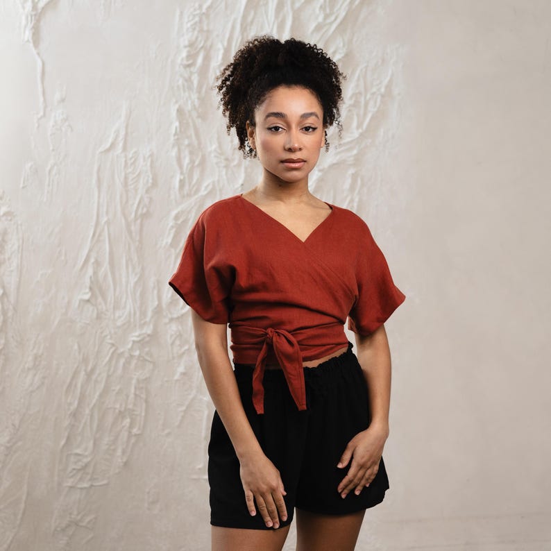 May include: A terracotta-coloured wrap top with short sleeves and a V-neck, tied at the waist. Worn with black shorts. The model is standing in front of a textured white wall.