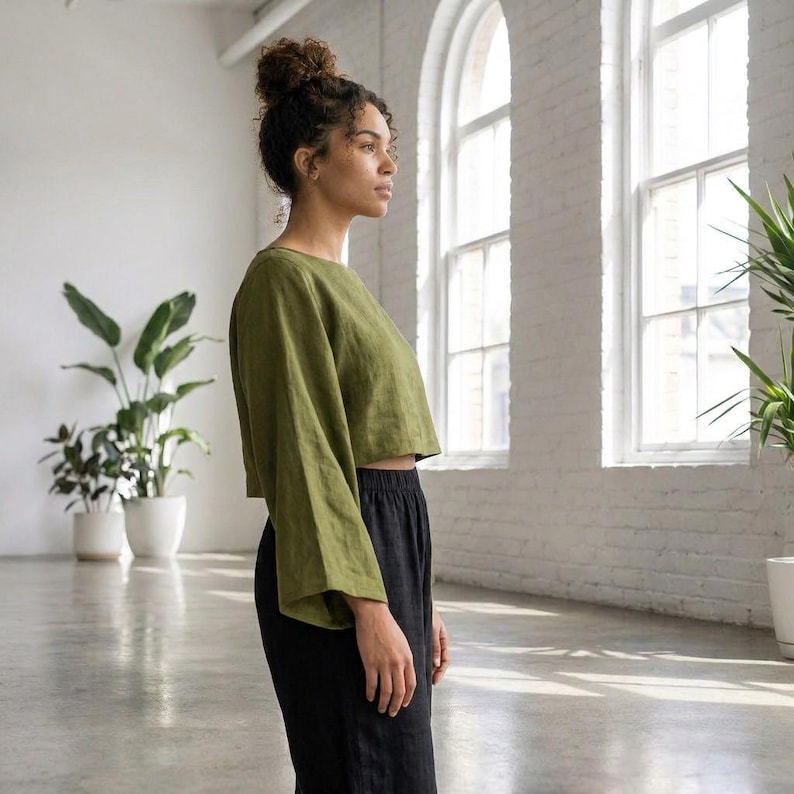 Puede incluir: Un top corto verde oliva de manga larga y pantalones negros. El top tiene un escote amplio y mangas sueltas. Los pantalones son de cintura alta y perneras anchas. El conjunto es de tejido ligero.