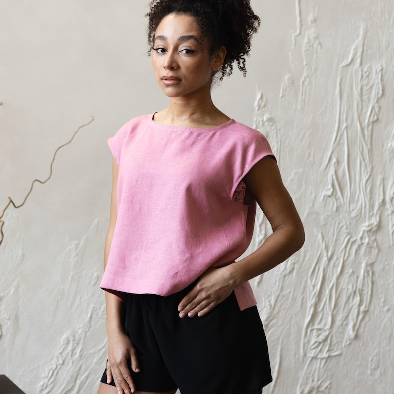 May include: A pink linen top with a square neckline and short sleeves. The top is paired with black shorts. The outfit is simple and casual, suitable for everyday wear. The fabric appears to be lightweight and breathable.