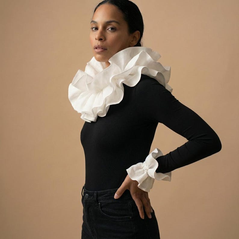Op de afbeelding: Een zwart shirt met lange mouwen met een grote witte kraag met ruches en bijpassende manchetten met ruches. Het model draagt een zwarte jeans. De achtergrond is effen beige. De top is een klassiek en elegant ontwerp.