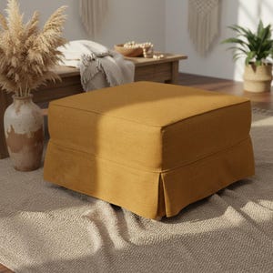 May include: A square, mustard-colored ottoman with a pleated skirt. The ottoman is made of a textured fabric and sits on a neutral-colored rug. The background includes a wooden table, a vase with dried plants, and a houseplant.