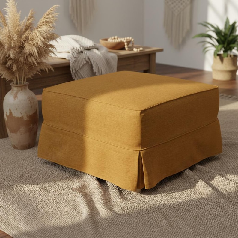 May include: A square, mustard-colored ottoman with a pleated skirt. The ottoman is made of a textured fabric and sits on a neutral-colored rug. The background includes a wooden table, a vase with dried plants, and a houseplant.