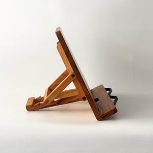 May include: A wooden book stand with a light brown and dark brown finish. The stand is adjustable and features a sloped surface to hold a book. The stand has a rectangular base and a black metal support.