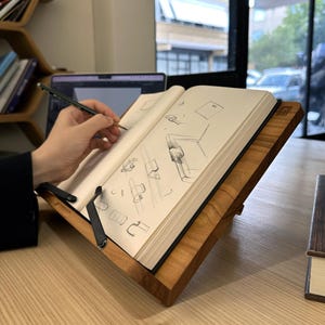 May include: A wooden book stand with a book open to sketches. The stand is made of light brown wood and has a black metal page holder. A person is drawing with a pencil on the open pages of the book.