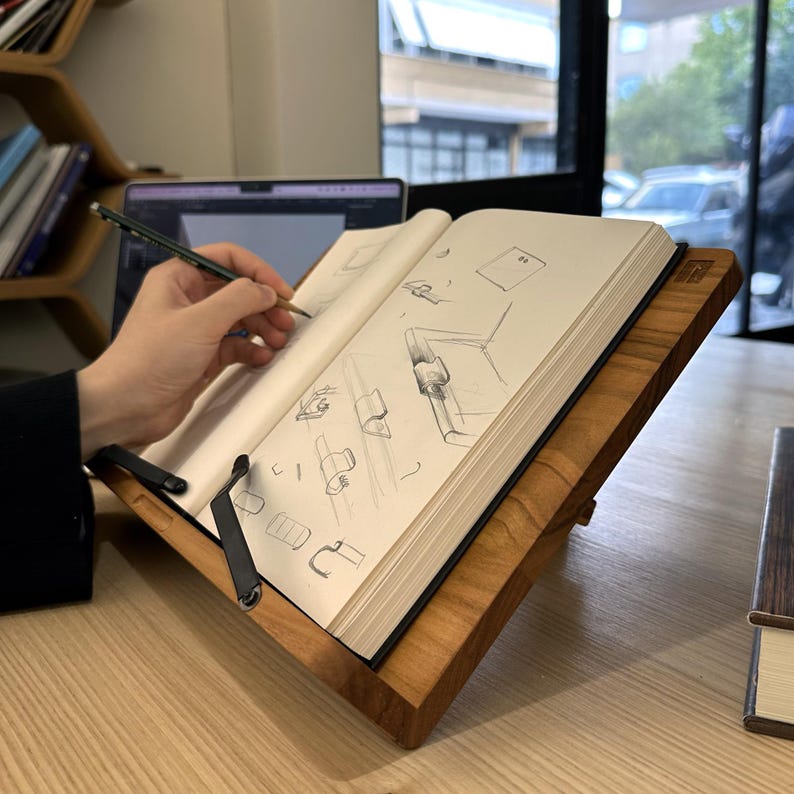 May include: A wooden book stand with a book open to sketches. The stand is made of light brown wood and has a black metal page holder. A person is drawing with a pencil on the open pages of the book.