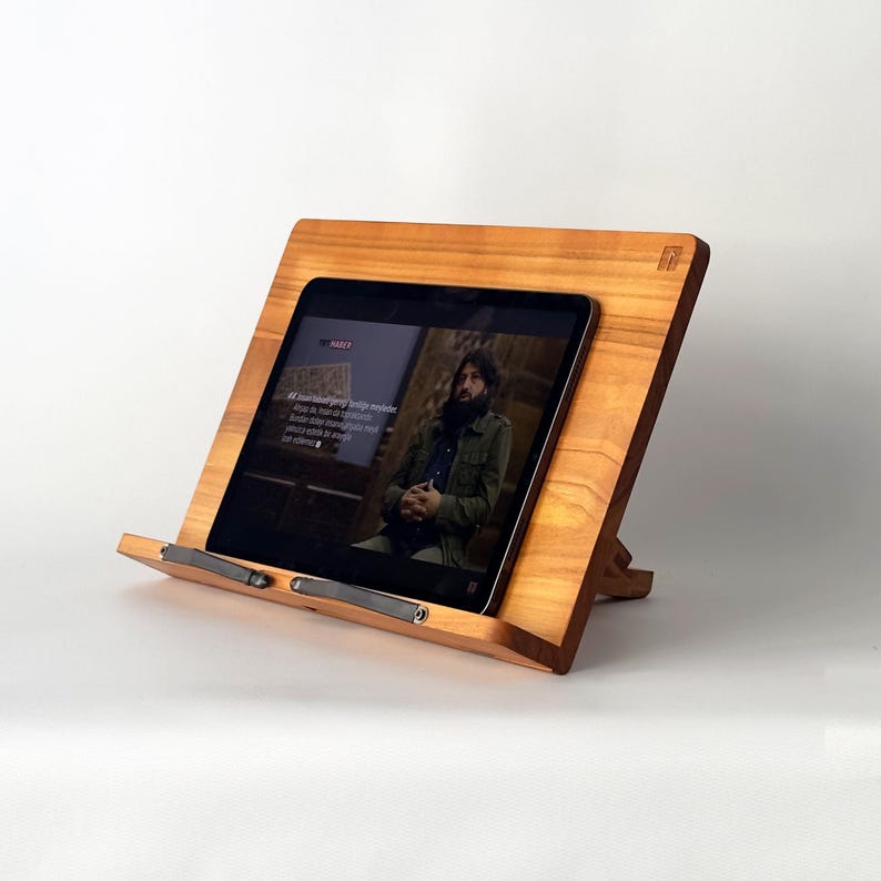 May include: A wooden tablet stand with a light brown finish. The stand is angled and holds a black tablet displaying a video. The stand has a small logo in the upper right corner and a metal bar to hold the tablet in place.