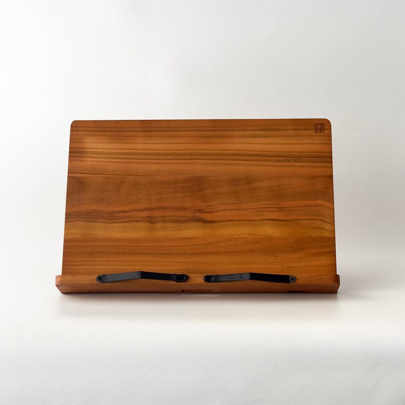 May include: A wooden book stand with a natural wood grain finish. The stand has a rectangular shape with a slightly angled back and a small ledge at the bottom. Two black metal clips hold the pages in place. A small square logo is visible on the top right.