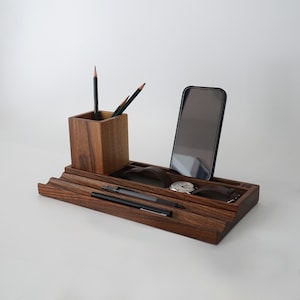 May include: A dark wood desk organizer with multiple compartments. It holds a smartphone, a wristwatch, pens, and pencils. The organizer has a pencil holder, a phone slot, and a watch holder. The wood grain is visible, and the overall design is minimalist.