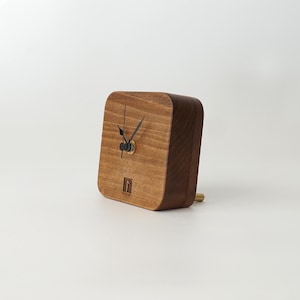 May include: A wooden clock with black hands and a brass center. The clock is square and has a light brown wood finish. The clock is sitting on a white surface.