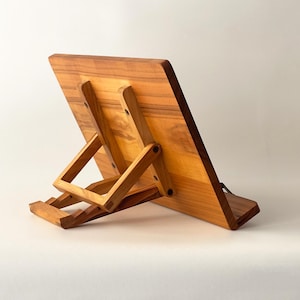 May include: A wooden book stand with a natural finish. The stand is angled, with a rectangular base and a hinged support system. The wood grain is visible, and the stand appears sturdy. The design is simple and functional, suitable for holding books or tablets.