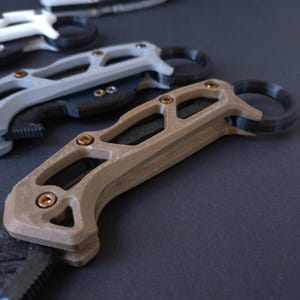 May include: A brown and black tactical knife with a skeletonized handle and a finger ring. The knife has a black blade and a brown handle with a textured surface.