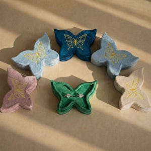Puede incluir: Una colección de joyeros en forma de mariposa, hechos de terciopelo, en tonos azul, rosa, verde y beige. Cada joyero tiene un diseño de mariposa bordada en oro. Un joyero verde contiene dos anillos.