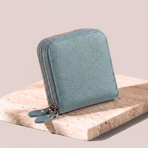 Closed sky blue pebble leather jewelry case sitting on a light stone surface, showing the rich grain texture and double gold-tone zippers.