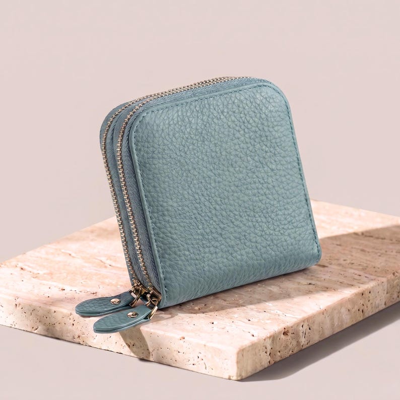 Closed sky blue pebble leather jewelry case sitting on a light stone surface, showing the rich grain texture and double gold-tone zippers.