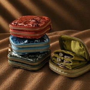 Op de afbeelding: Een stapel kleine, vierkante fluwelen sieradendoosjes in tinten roest, blauw en groen, met gouden ritsen. Eén doosje is open en toont meerdere ringen. De doosjes liggen op een bruin oppervlak.