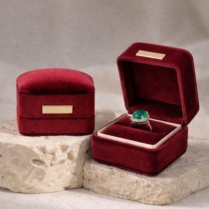 Two royal burgundy velvet jewelry boxes on a textured surface. The closed box features a signature gold bar, while the open box holds a luxury gemstone ring, ideal for a classic wedding ceremony.