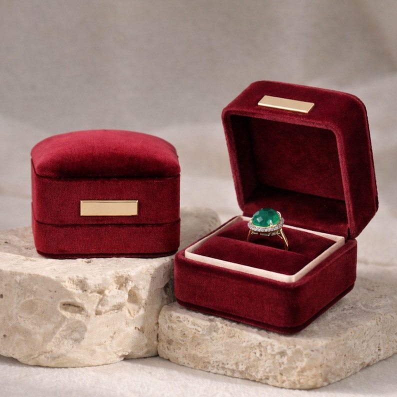 Two royal burgundy velvet jewelry boxes on a textured surface. The closed box features a signature gold bar, while the open box holds a luxury gemstone ring, ideal for a classic wedding ceremony.