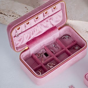 May include: Open pink velvet jewellery box with gold zip and compartments. The interior features ring slots, earring holders, and necklace hooks. Various rings, earrings, and necklaces are displayed inside the box, showcasing its storage capacity.
