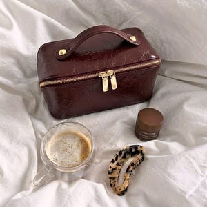 Könnte beinhalten: Eine weinrote Kosmetiktasche aus Kunstleder mit goldenem Reißverschluss und Griff. Eine Tasse Kaffee, ein kleines Glas und eine Haarklammer mit Schildpattmuster sind ebenfalls vorhanden.