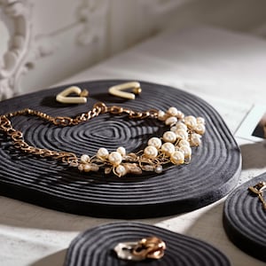 May include: A gold-toned necklace with pearl accents and matching earrings rest on a black, textured surface. The necklace features a delicate chain and floral-inspired details. The earrings are a cream color. Additional jewelry is visible.