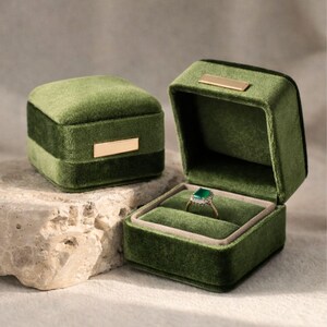 A set of two olive green velvet ring holders: one closed showing the sleek gold accent, and one open displaying a sparkling emerald engagement ring. Professional wedding flat lay prop.