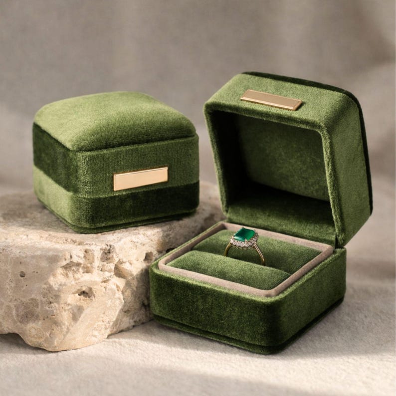 A set of two olive green velvet ring holders: one closed showing the sleek gold accent, and one open displaying a sparkling emerald engagement ring. Professional wedding flat lay prop.