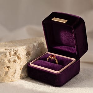 A rich royal purple velvet ring box open with an antique gold ruby ring inside. The deep violet tones and plush velvet interior emphasize a high-end, majestic aesthetic.