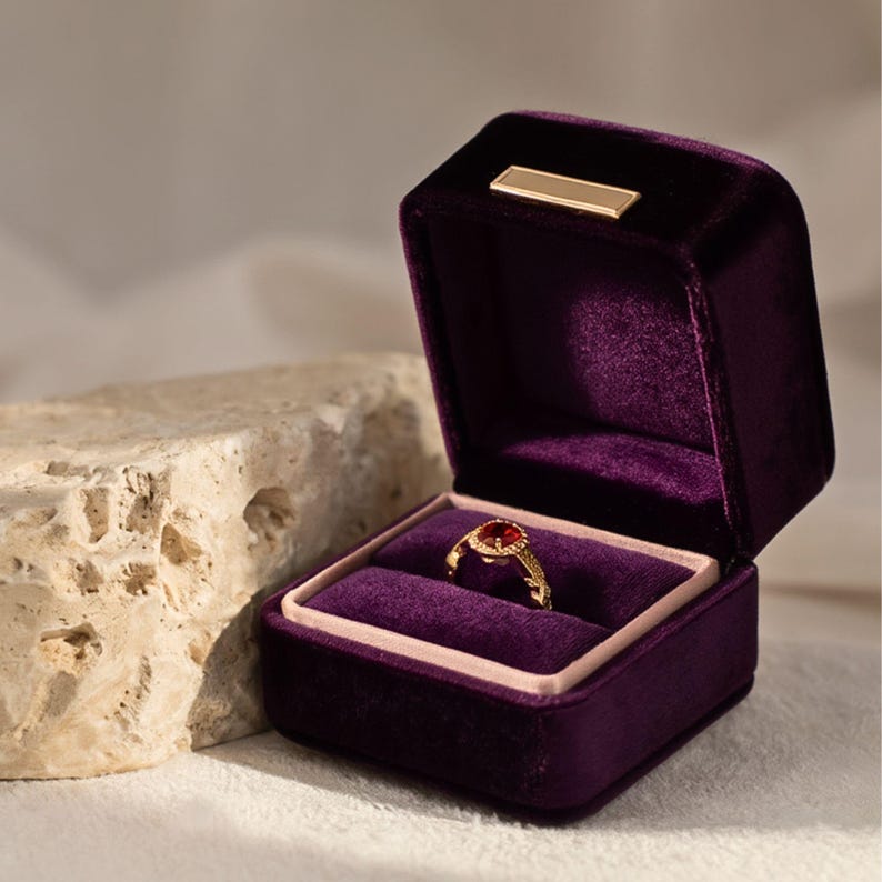 A rich royal purple velvet ring box open with an antique gold ruby ring inside. The deep violet tones and plush velvet interior emphasize a high-end, majestic aesthetic.