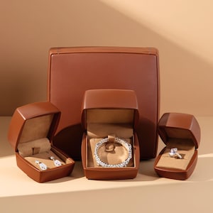 Könnte beinhalten: Eine Sammlung brauner Leder-Schmuckkästchen, die jeweils geöffnet sind, um ein anderes Schmuckstück zu präsentieren. Der Schmuck umfasst ein Paar Ohrringe, ein Armband und einen Ring. Die Kästchen sind vor einem größeren, passenden Lederetui angeordnet.