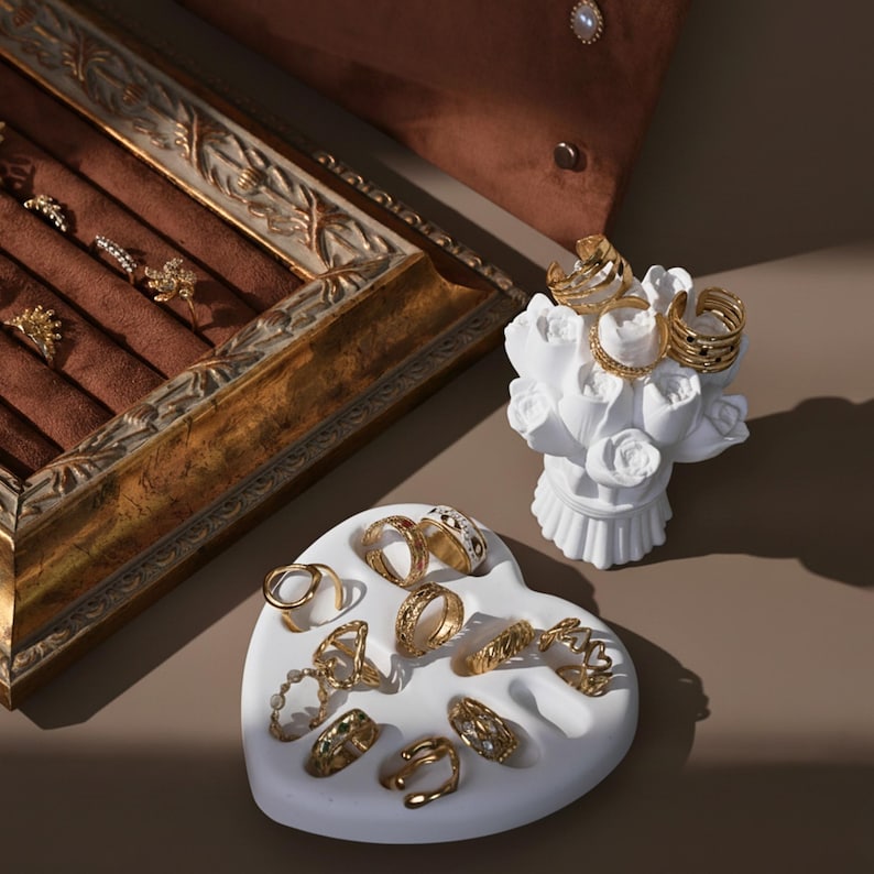 An aesthetic collection of jewelry props, including a vintage frame, a heart-shaped dish, and a rose bouquet holder, all displaying gold rings.