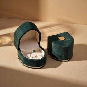 Può includere: Un anello in argento con una piccola pietra preziosa rotonda di un verde vibrante si trova all'interno di un portagioie in velluto verde scuro aperto. Una seconda scatola in velluto verde scuro chiusa con un piccolo dettaglio in metallo dorato.
