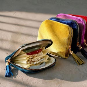 Op de afbeelding: Een verzameling kleurrijke fluwelen sieradenetuis met gouden ritsen en kwastjes. Eén etui is open en toont sieraden op crèmekleurige stof. De etuis zijn in tinten van teal, geel, blauw, paars en rood.