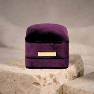 A closed minimalist purple velvet jewelry case sitting on a natural rock. The focus is on the luxurious fabric texture and the polished gold-tone rectangular accent plate.