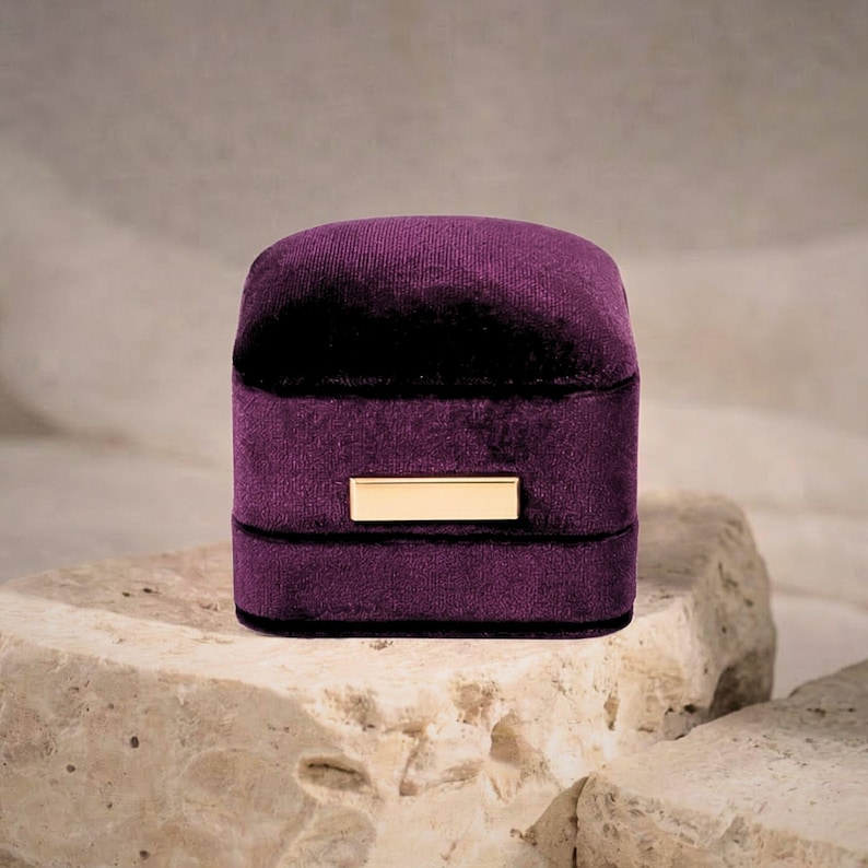 A closed minimalist purple velvet jewelry case sitting on a natural rock. The focus is on the luxurious fabric texture and the polished gold-tone rectangular accent plate.