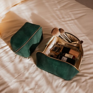Puede incluir: Dos neceseres de maquillaje de pana verde con cremalleras doradas sobre una cama blanca. Una bolsa está abierta, revelando brochas de maquillaje y varios productos cosméticos. La otra bolsa está cerrada.