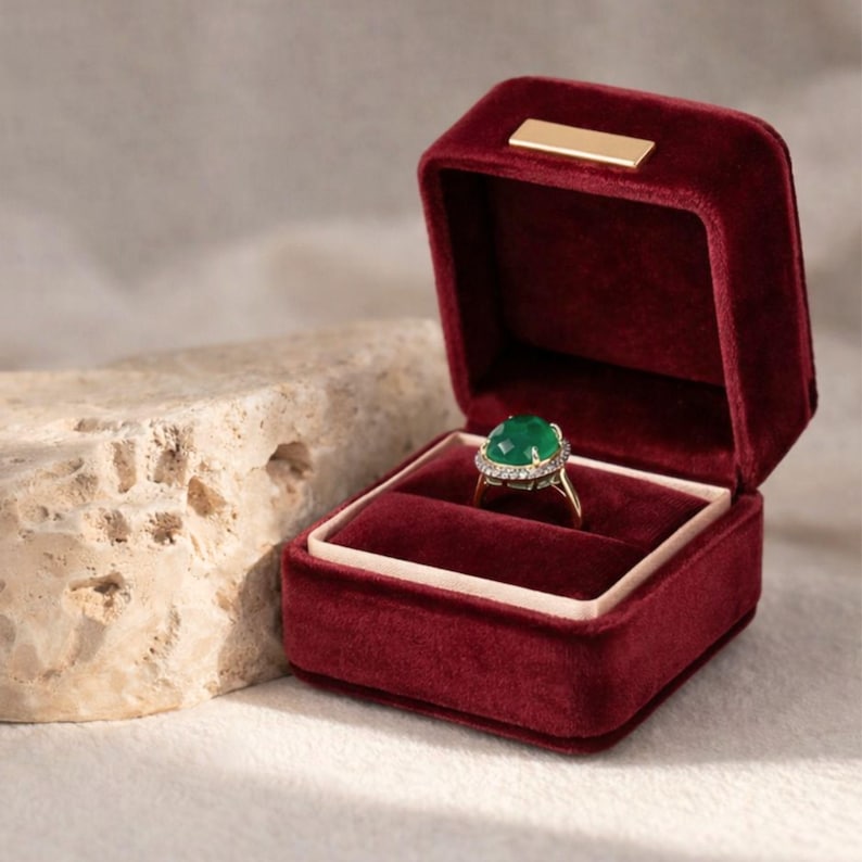Deep burgundy red velvet ring box open to reveal a vintage-style green gemstone ring. The rich wine-colored fabric contrasts beautifully with the natural stone background.