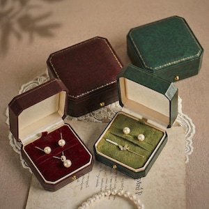 Puede incluir: Cajas de joyería burdeos y verdes, con detalles dorados. Una caja está abierta, revelando un collar y pendientes de perlas. Otra caja muestra un collar con colgante y pendientes de perlas. Las cajas están sobre una superficie color crema.