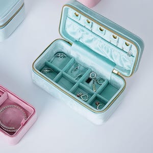 Puede incluir: Caja de joyería de terciopelo azul claro abierta, con cremallera dorada y compartimentos para anillos, pendientes y collares. El interior está forrado con una tela de satén azul claro a juego. Varias joyas son visibles dentro de la caja.