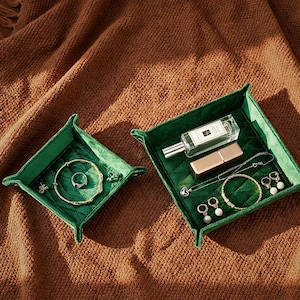 Könnte beinhalten: Zwei grüne Samttabletts mit Schmuck und Schönheitsprodukten. Ein Tablett enthält ein goldenes Armband, einen Ring und Ohrringe. Das andere Tablett enthält eine Parfümflasche, Lippenstift, Halskette, Armband und Ohrringe.