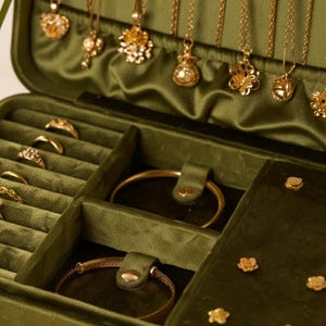Könnte beinhalten: Eine grüne Samt-Schmuckschatulle gefüllt mit goldenen Halsketten, Ringen und Armbändern. Die Halsketten hängen von oben, während Ringe und Armbänder in Fächern angeordnet sind. Die Schatulle ist geöffnet und zeigt den Schmuck.