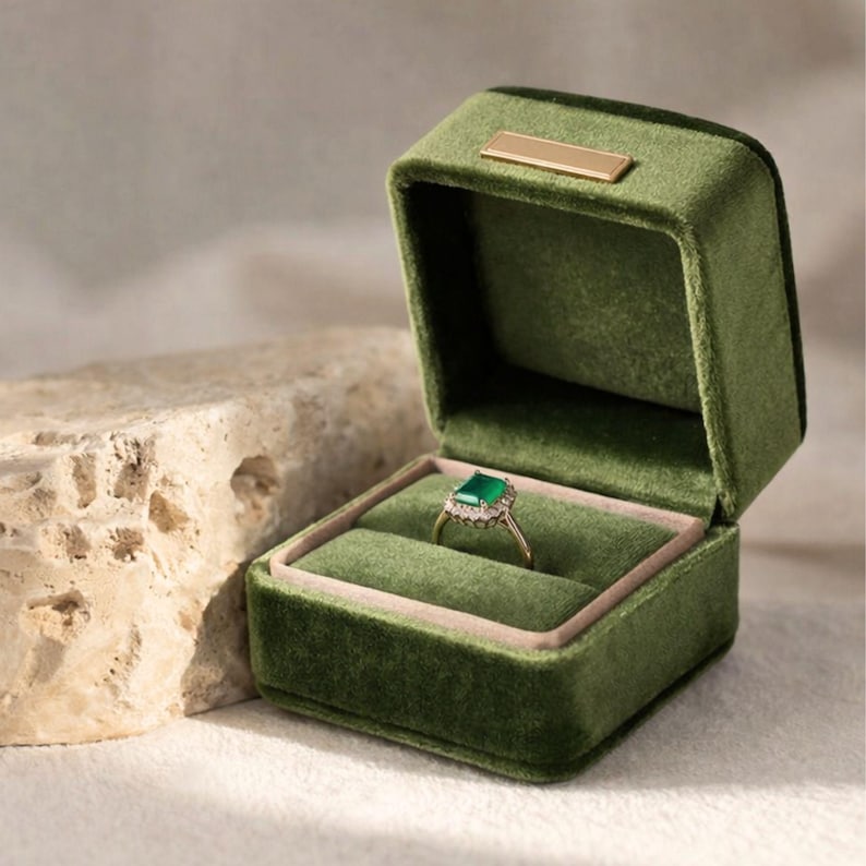 An open moss green velvet ring box with a minimalist gold nameplate, showcasing an emerald-cut green gemstone ring. The box sits on a natural travertine stone under soft, warm lighting.
