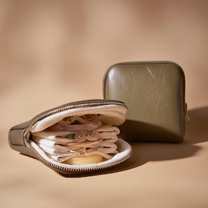 Puede incluir: Estuche de viaje para joyas verde oliva abierto, con cremallera dorada, que revela múltiples pulseras y anillos. Una segunda funda verde oliva cerrada se encuentra detrás de la funda abierta. El interior de la funda abierta está forrado con tela blanca.