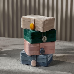 Op de afbeelding: Een stapel van vier rechthoekige fluwelen sieradendoosjes in beige, smaragdgroen, poederroze en blauwgrijs. Elk doosje heeft een bijpassende band en een decoratieve stenen sluiting. De doosjes zijn op elkaar gestapeld.