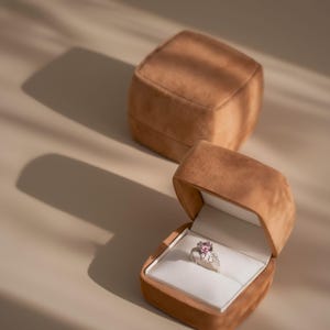 Puede incluir: Un anillo de plata con una piedra preciosa rosa y pequeñas piedras blancas se encuentra dentro de un joyero de terciopelo marrón abierto. Otro joyero cerrado está en el fondo. Las cajas son cuadradas con bordes redondeados y el interior es blanco.