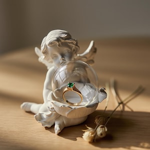 Peut inclure: Une statuette de chérubin blanc tenant un globe de verre transparent contenant une bague en or avec une pierre précieuse verte. La bague est au centre, sur un fond neutre. Des fleurs séchées sont au premier plan.