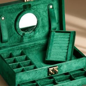 Op de afbeelding: Open smaragdgroene fluwelen sieradendoos met een ingebouwde spiegel en meerdere compartimenten. De doos heeft een ringhouder en verschillende secties voor het opbergen van kettingen, armbanden en oorbellen. De doos is beveiligd met een goudkleurige sluiting.