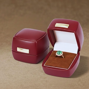May include: Two claret-coloured ring boxes, one open and one closed, with gold-coloured accents. The open box displays a gold ring with a green gemstone and a halo of smaller stones, set on a brown velvet interior.
