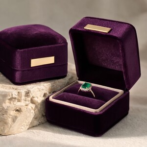 Pair of luxury violet purple velvet ring boxes. The open box showcases a diamond and emerald ring, creating a sophisticated look for jewelry branding or wedding detail shots.